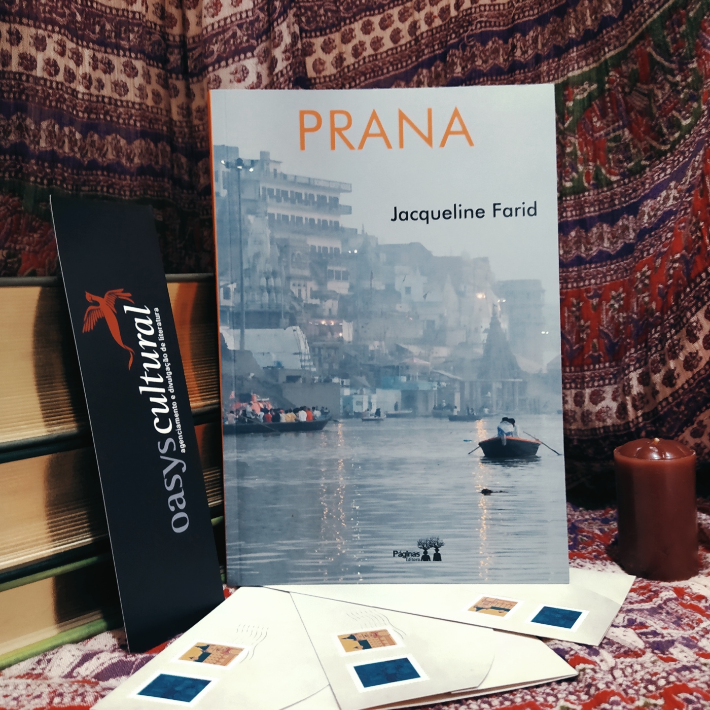 Livro Prana apoiado em uma pilha de livros, com um marcador da Oasys Cultural à esquerda e uma vela à direita. Abaixo, há três envelopes de cartas com selos. Ao fundo, há um tecido com tema que remete à Índia.