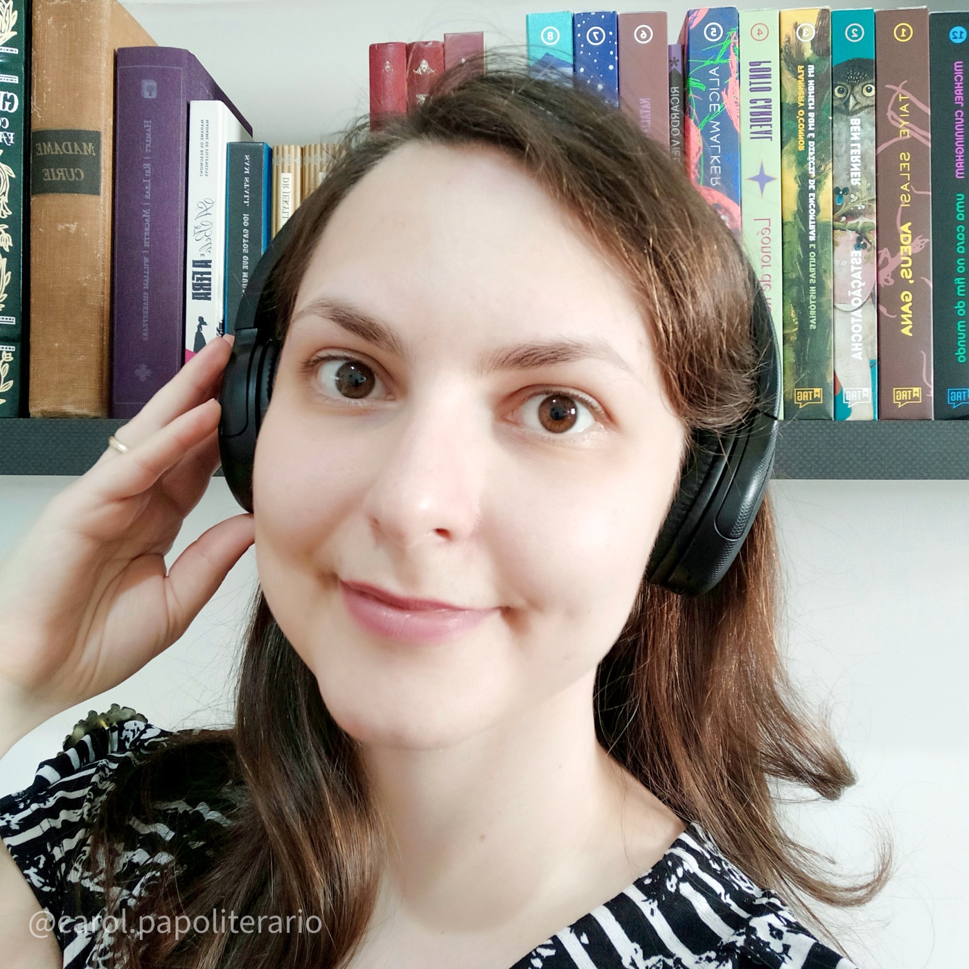 Foto de uma moça sorrindo, com um fone de ouvido estilo headphone, e cabelos soltos. No fundo, há uma estante de livros.
