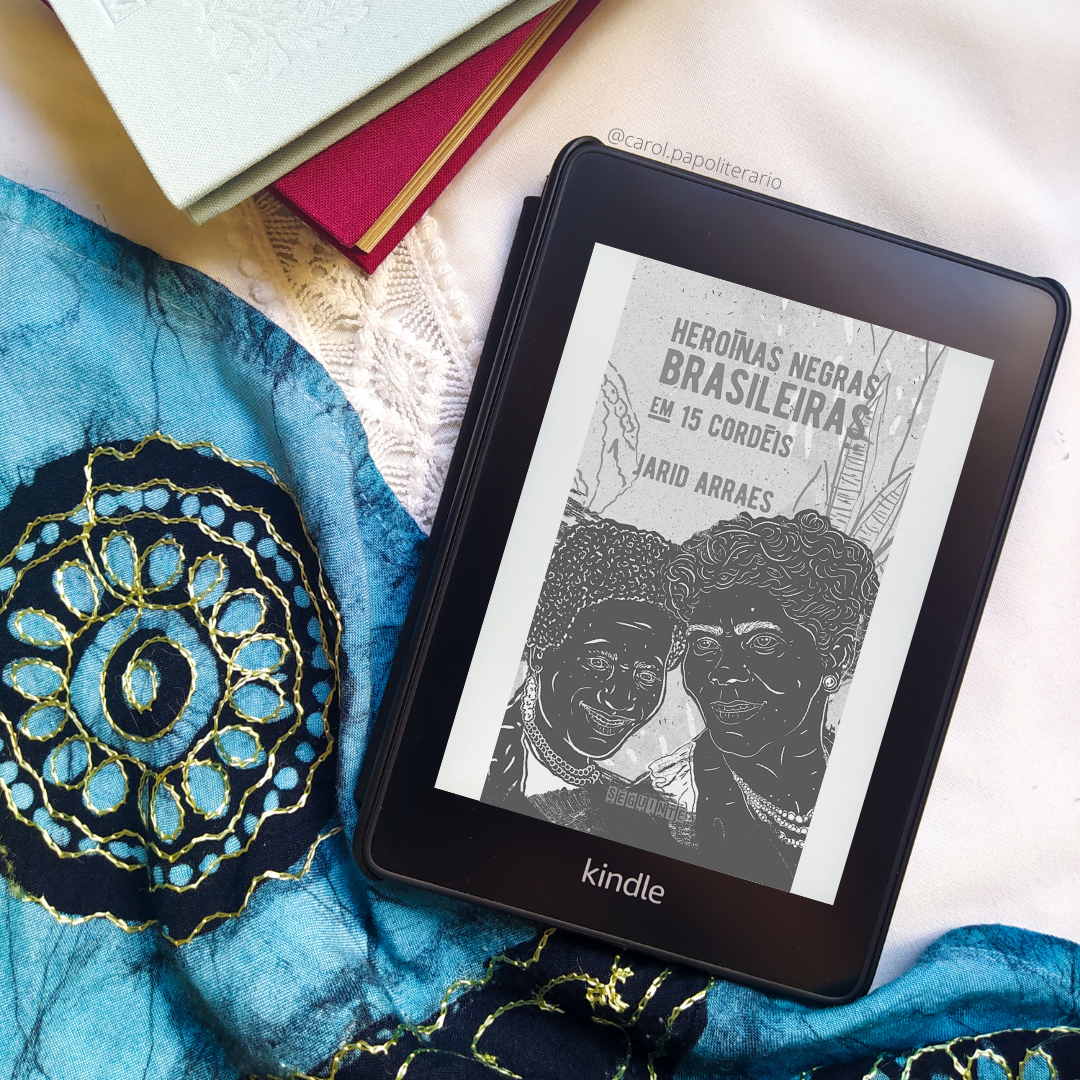 Kindle com a capa do livro Heroínas Negras Brasileiras em 15 cordéis. Há uma pilha de livros com capa de tecido no canto superior esquerdo da imagem. Há um tecido com motivos indianos na metade diagonal inferior esquerda da foto.